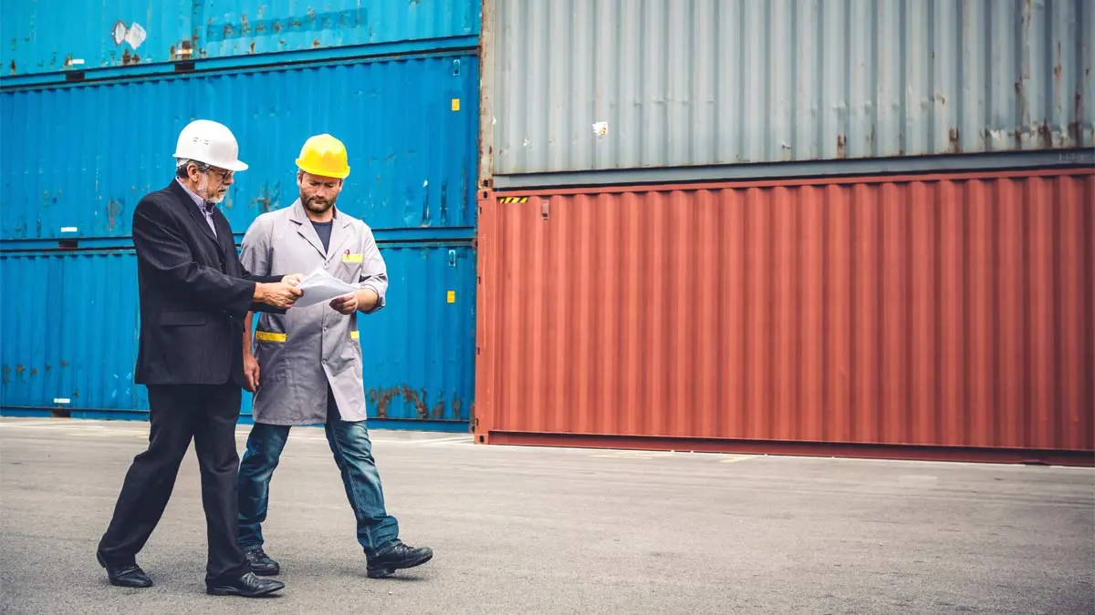 Two men in hard hats walking through an aisle of freight containers, holding a document